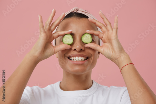 Head and shoulders portrait of Mixed-Race young woman holding cucumber slices and smiling happily at camera while posing against pink background