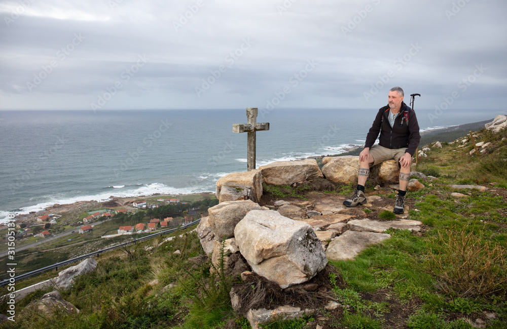 A hiker takes a break and sits high on a mountain on the Atlantic coast in Galicia in northern Spain. A stone cross stands in front of him. It is cloudy and you can see the coastline.