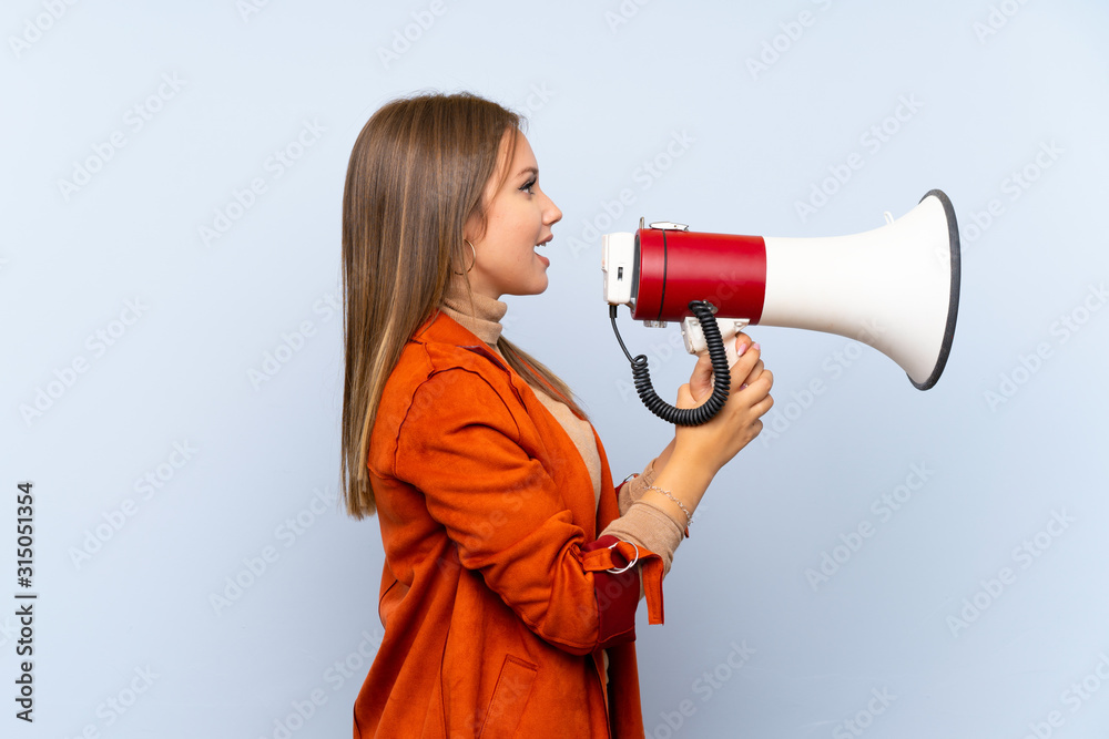 Teenager girl with coat over isolated blue background shouting through a megaphone Teenager girl with coat over isolated blue background shouting through a megaphone