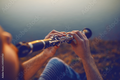 Close up of handsome caucasian bearded blond man sitting on cliff and playing clarinet.