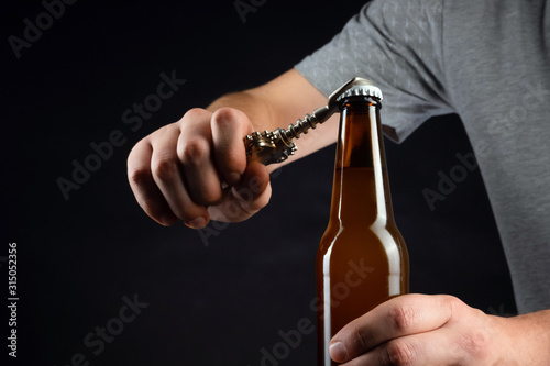 Men opening cold bottle of beer with cap on black background. Hands cracking refrigerated wheat or lager beer with an opener on dark background