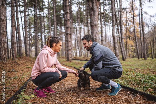 Happy young couple in sportswear crouching on trail in woods, looking at each other and petting stray dog. Break after running. Healthy lifestyle concept.