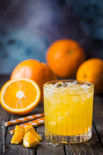 Glass jar of fresh orange juice with ice and fresh fruits on dark table.