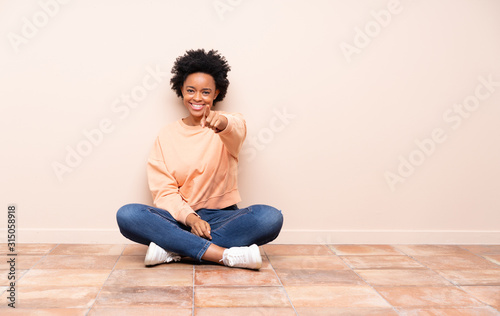 African american woman sitting on the floor points finger at you with a confident expression © luismolinero