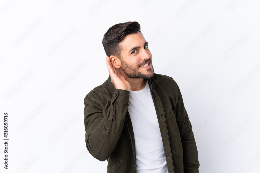 Young handsome man with beard over isolated white background listening to something by putting hand on the ear Young handsome man with beard over isolated white background listening to something by putting hand on the ear