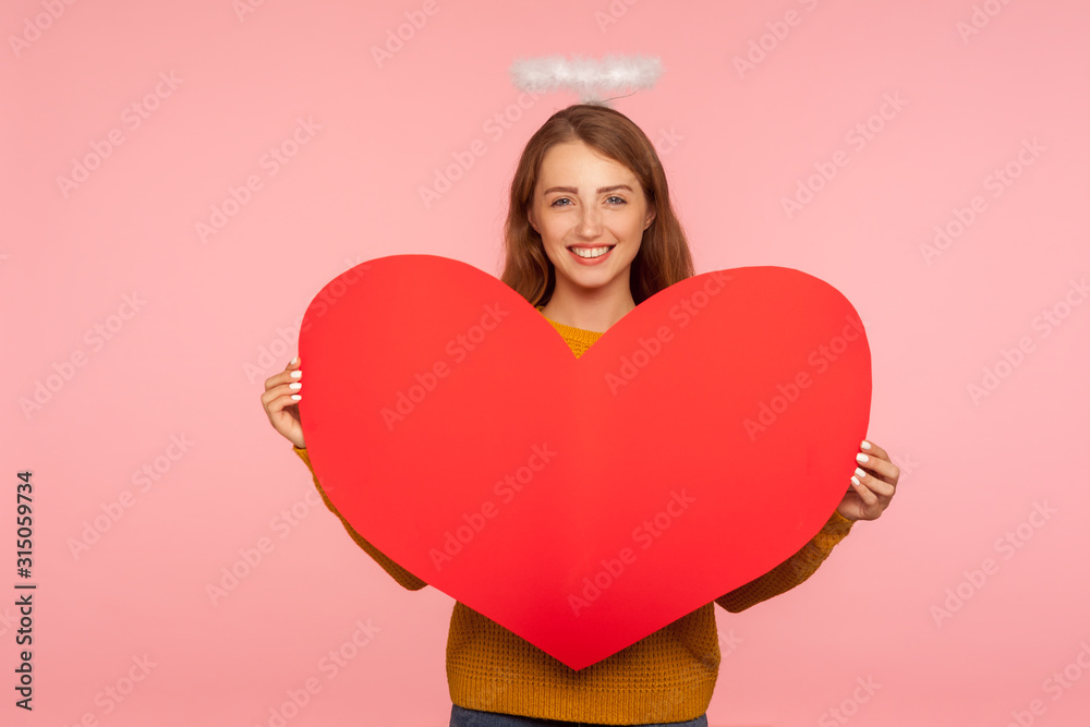 Angel on Valentine's day. Portrait of lovely ginger girl with halo over head smiling at camera and holding big red heart, symbol of love and affection, romantic feelings. studio shot, pink background Angel on Valentine's day. Portrait of lovely ginger girl with halo over head smiling at camera and holding big red heart, symbol of love and affection, romantic feelings. studio shot, pink background