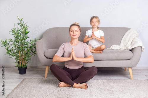 Lotus posture. Young mother and little daughter with closed eyes and prayer gesture practicing yoga together at home, doing exercise breath technique, meditating in room, mindfulness and harmony