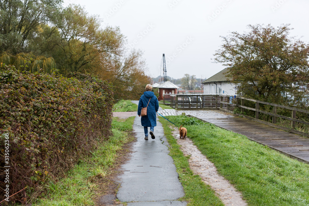 A middle aged woman walking her small dog along a river path on wet and rainy day