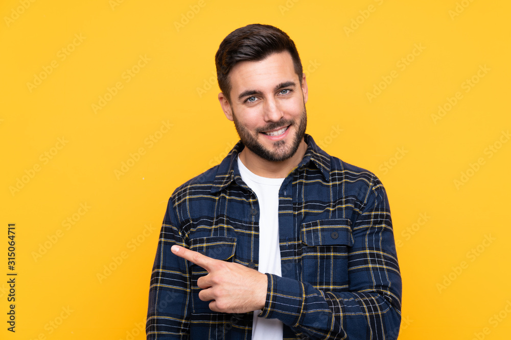 Young handsome man with beard over isolated yellow background pointing to the side to present a product Young handsome man with beard over isolated yellow background pointing to the side to present a product