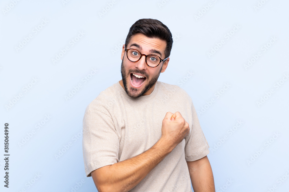 Young handsome man with beard over isolated blue background celebrating a victory Young handsome man with beard over isolated blue background celebrating a victory