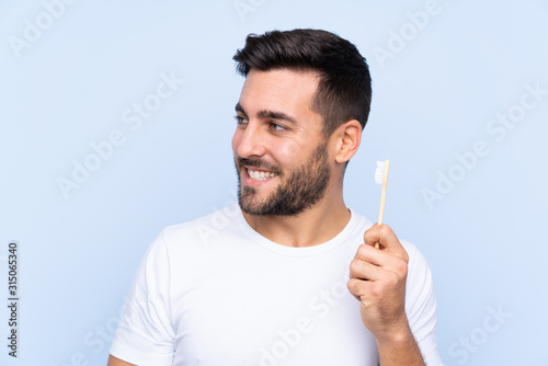 Young handsome man with beard brushing his teeth over isolated background looking side