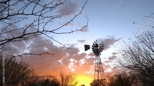 Windpump against a dramatic sunset with orange clouds on a sheep farm near Keetmanshoop, Namibia. Thorntree branches in the foreground. Stable wide shot. photo