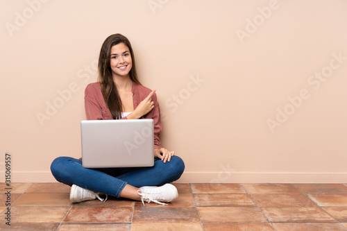 Teenager student girl sitting on the floor with a laptop pointing to the side to present a product