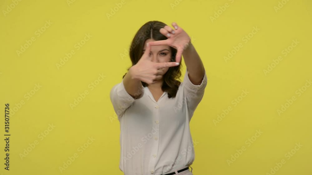 Attentive woman with brunette hair in blouse looking through frame made of fingers , focusing on camera with curious expression, capturing photo. indoor studio shot isolated on yellow background Attentive woman with brunette hair in blouse looking through frame made of fingers , focusing on camera with curious expression, capturing photo. indoor studio shot isolated on yellow background