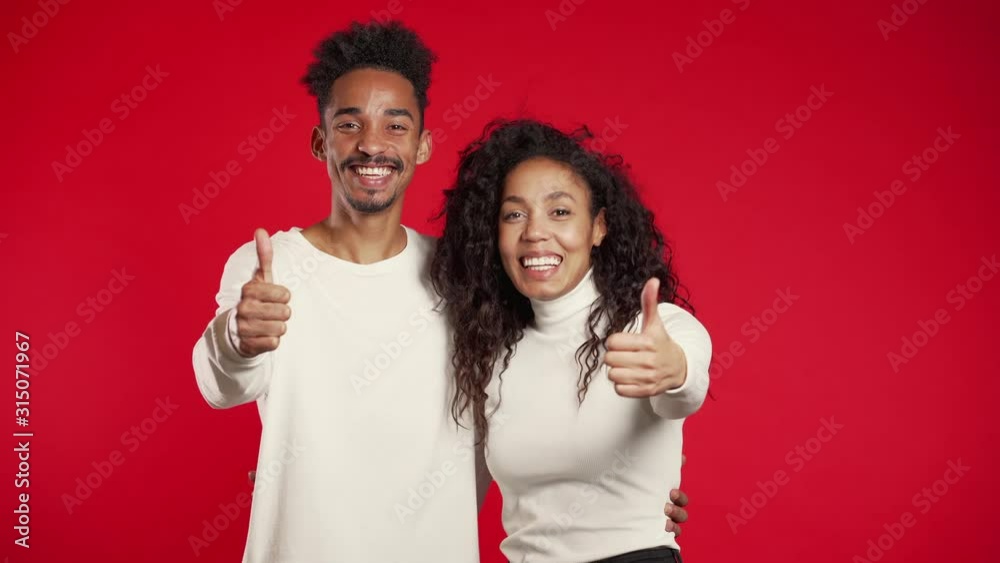 Young african american couple making thumbs up sign over red background. Winner. Success. Positive girl and man smiles to camera. Body language. Young african american couple making thumbs up sign over red background. Winner. Success. Positive girl and man smiles to camera. Body language.