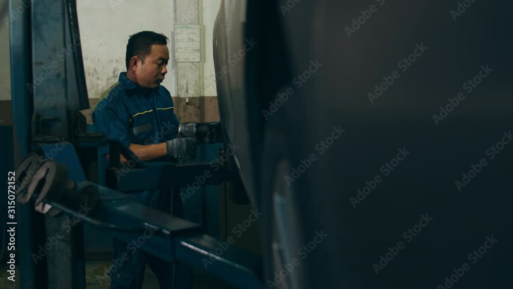 Professional car mechanic changing a car tire on lifted automobile at repair service station. Wheel alignment work at workshop night. Skillful Asian guy in uniform fixing car. 4k slow motion shot. Professional car mechanic changing a car tire on lifted automobile at repair service station. Wheel alignment work at workshop night. Skillful Asian guy in uniform fixing car. 4k slow motion shot.