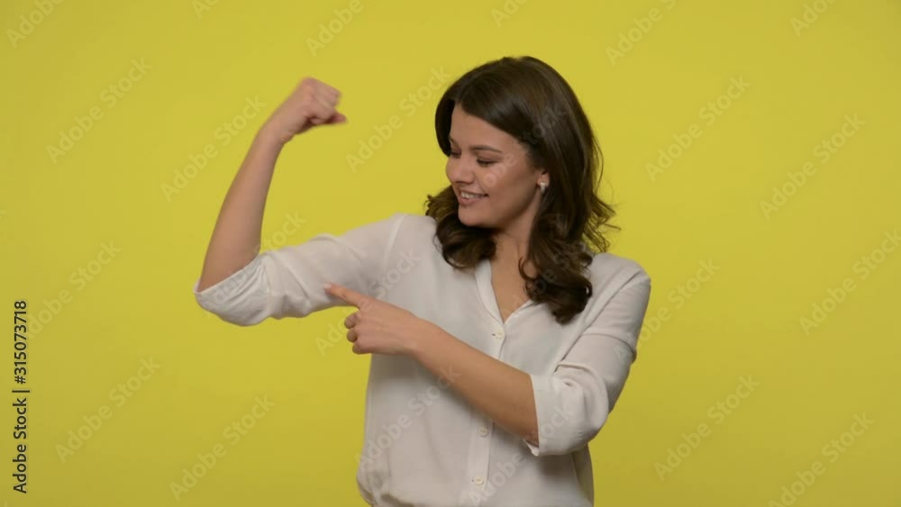 I'm strong! Confident positive businesswoman with brunette hair in blouse pointing biceps, showing strength, feeling powerful and successful, feminism. indoor studio shot isolated on yellow background I'm strong! Confident positive businesswoman with brunette hair in blouse pointing biceps, showing strength, feeling powerful and successful, feminism. indoor studio shot isolated on yellow background