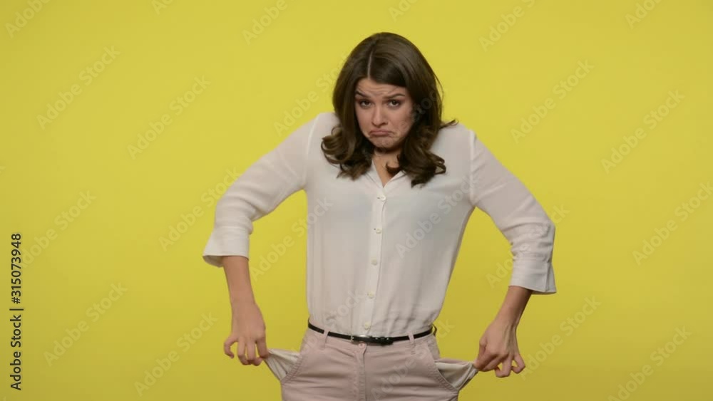 Vexed woman with brunette hair in blouse turning out pockets and looking at camera with frustrated expression, showing no money, going bankrupt. indoor studio shot isolated on yellow background