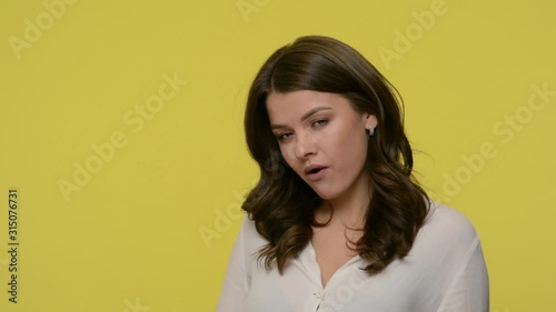 Nice woman with brunette hair in blouse looking with suspicious at camera and chewing gum, giving a smile with arrogant disrespectful look, bad manner habit. studio shot isolated on yellow background
