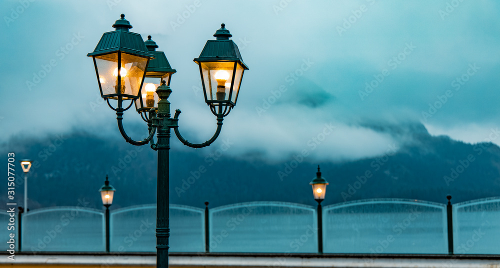 moody street lantern yellow illumination autumn time rainy weather day waterfront European village sidewalk district with fence and foggy dramatic mountain landscape background with empty copy space