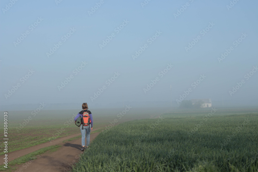 Woman traveler with backpack returns home in beautifyl atmospheric misty morning a house among fields in the background Woman traveler with backpack returns home in beautifyl atmospheric misty morning a house among fields in the background