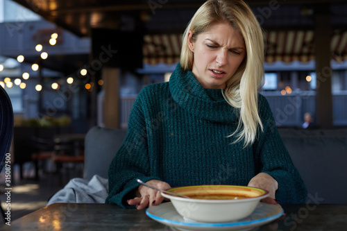Blonde Caucasian woman in green pullover demonstrates disgust twisting face with negative reaction while trying to eat some smelly soup from a plate on a table