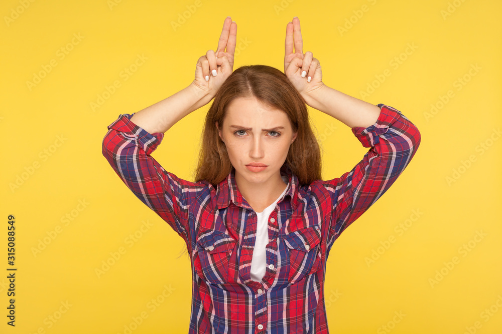 I'm a cow. Portrait of angry menacing ginger girl in checkered shirt showing bull horns on head, threatening to attack, danger and aggression concept. indoor studio shot isolated on yellow background I'm a cow. Portrait of angry menacing ginger girl in checkered shirt showing bull horns on head, threatening to attack, danger and aggression concept. indoor studio shot isolated on yellow background