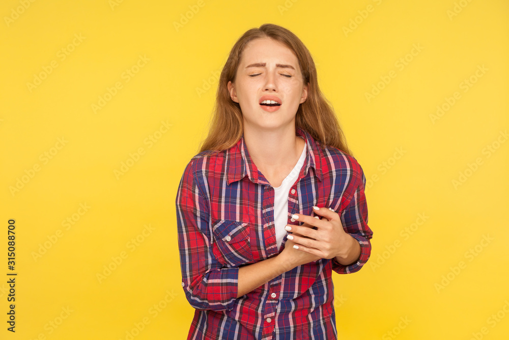 Portrait of young ginger woman in checkered shirt shouting and grimacing from pain, clasping chest, suffering heart ache or breast pain, cardiac problems. studio shot isolated on yellow background Portrait of young ginger woman in checkered shirt shouting and grimacing from pain, clasping chest, suffering heart ache or breast pain, cardiac problems. studio shot isolated on yellow background