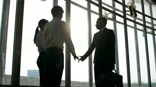 Medium shot of black businessman in suit walking towards male and female business partners and exchanging handshake in greeting before Panoramic windows on sunny day photo