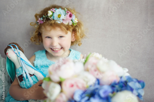 Portrait of 5 years old curly blonde laughing girl in blue dress and wreath with rabbit toy in hands. Waiting for spring small beautiful girl with big bouquet of artificial flowers