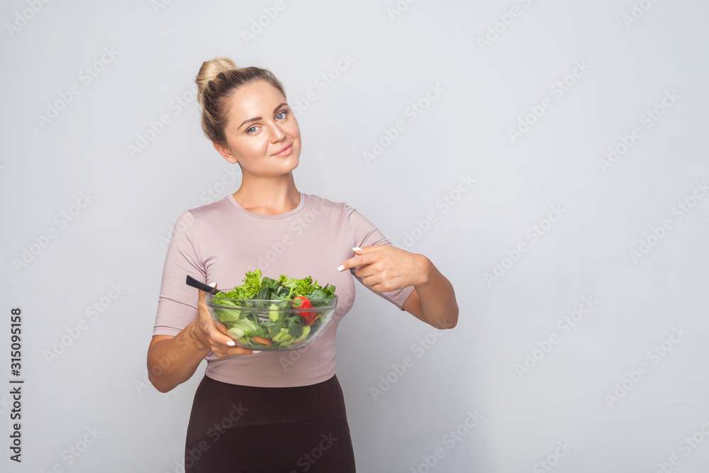 Portrait of satisfied fit woman with hair bun pointing at bawl of vegetable salad and looking at camera, vegetarian diet, healthy food, empty copy space for advertising. studio shot, grey background Portrait of satisfied fit woman with hair bun pointing at bawl of vegetable salad and looking at camera, vegetarian diet, healthy food, empty copy space for advertising. studio shot, grey background