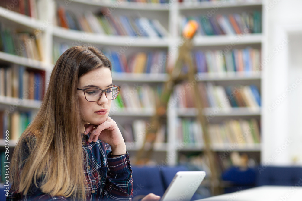 Girl in glasses working with tablet sitting in library. Student prefers to work with a tablet than books Girl in glasses working with tablet sitting in library. Student prefers to work with a tablet than books