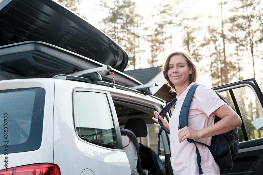 Contented woman with a backpack is going on a trip, she is standing near an open car and trunk, and is preparing to load things.