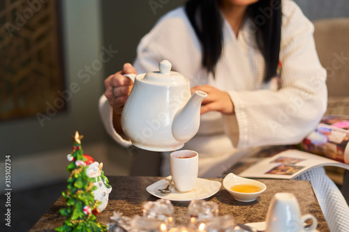 Relaxation and tea party in the spa, a girl in a bathrobe pours tea from a white teapot, there is a mug with tea and honey on the table