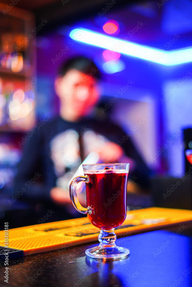 A transparent glass with mulled wine stands on a table on a blue background, behind a man stands on a blurred background, bright colors, side view