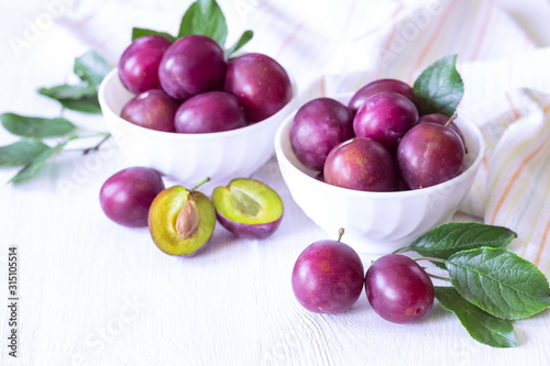 ripe purple plums in white cups on a white background. whole plums and half plums in bowls and on the table close-up.