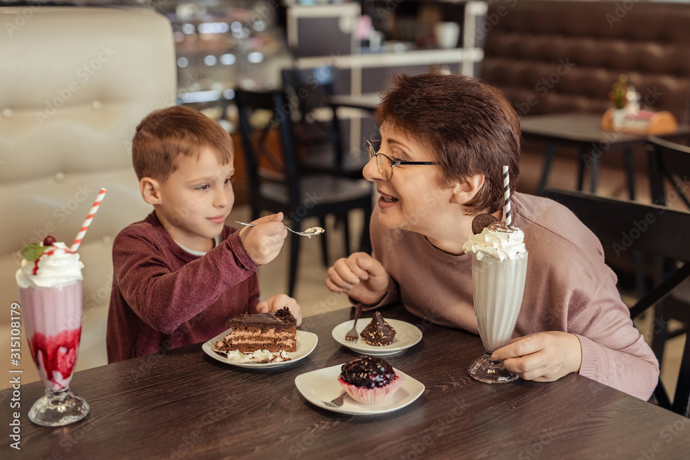 A happy grandmother with glasses and her grandson spend 7 years together in a cafe. they eat sweet cakes and milkshakes.