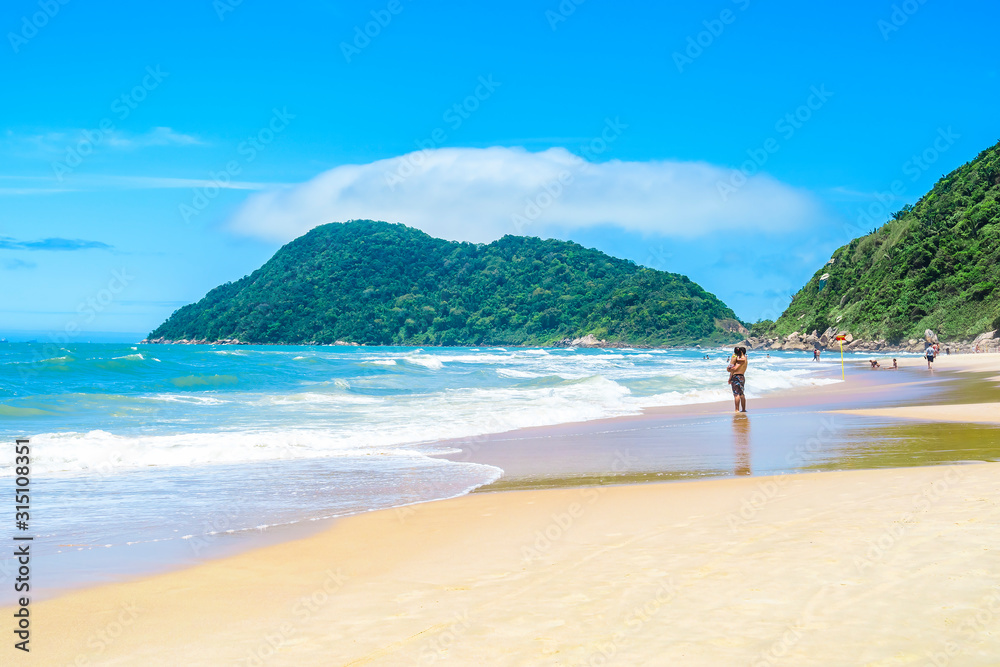 Sunbathers at a beautiful Brazilian beach of the Paulista coast on a sunny day. The sea, some waves, the sand and a hill on background. Photo at Praia do Tombo beach - Guaruja SP Brazil. Sunbathers at a beautiful Brazilian beach of the Paulista coast on a sunny day. The sea, some waves, the sand and a hill on background. Photo at Praia do Tombo beach - Guaruja SP Brazil.