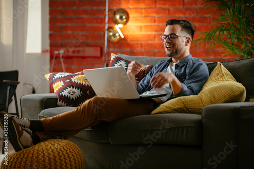 Young man sitting on sofa at home. Businessman celebrating. Attractive man using laptop.