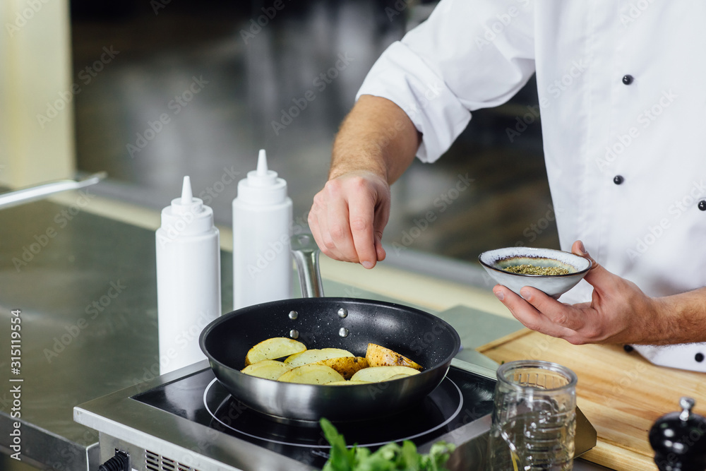 Male chef cooking fresh potato, sauce in restaurant kitchen. Cook at work during dinner service. Italian style cuisine, lifestyle and healthy food concept. Focus on his hand.