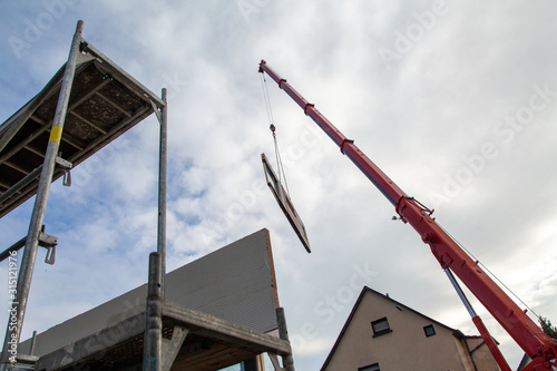 Floating component on a prefabricated house on the boom of a mobile crane with parts of a scaffolding in the foreground and residential building in the background