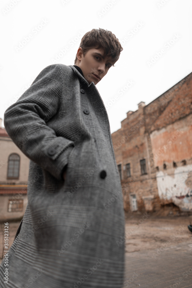Foto Stock Pretty attractive young man with brown hair in a long trendy  gray classic checkered coat is standing outside in the city on an autumn  day. Fashionable handsome guy outdoors. Trendy