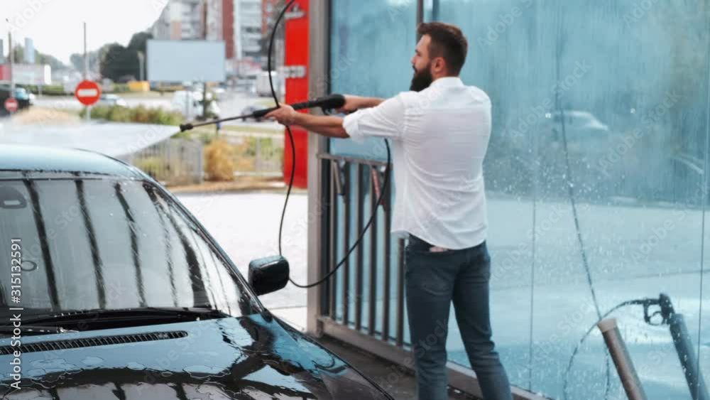 A driver has a beard. A car is black. The man is using water to wash the car. A driver has a beard. A car is black. The man is using water to wash the car.