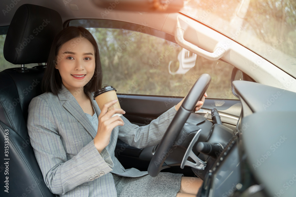 Asian female driver smiling and drinking coffee in the car, Beautiful girl  holding an eco paper coffee cup,looking to camera while driving her car, happy life transport in city