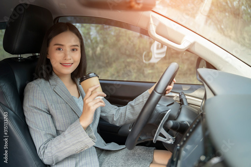 Asian female driver smiling and drinking coffee in the car, Beautiful girl holding an eco paper coffee cup,looking to camera while driving her car, happy life transport in city