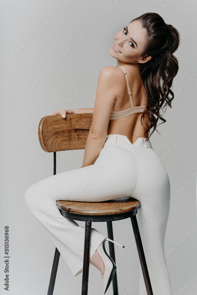 Back view of fashionable sexy woman in bra and white trousers leans at  chair, poses for making erotic photo, models in studio, has sensual smile,  minimal makeup, isolated over grey background Stock