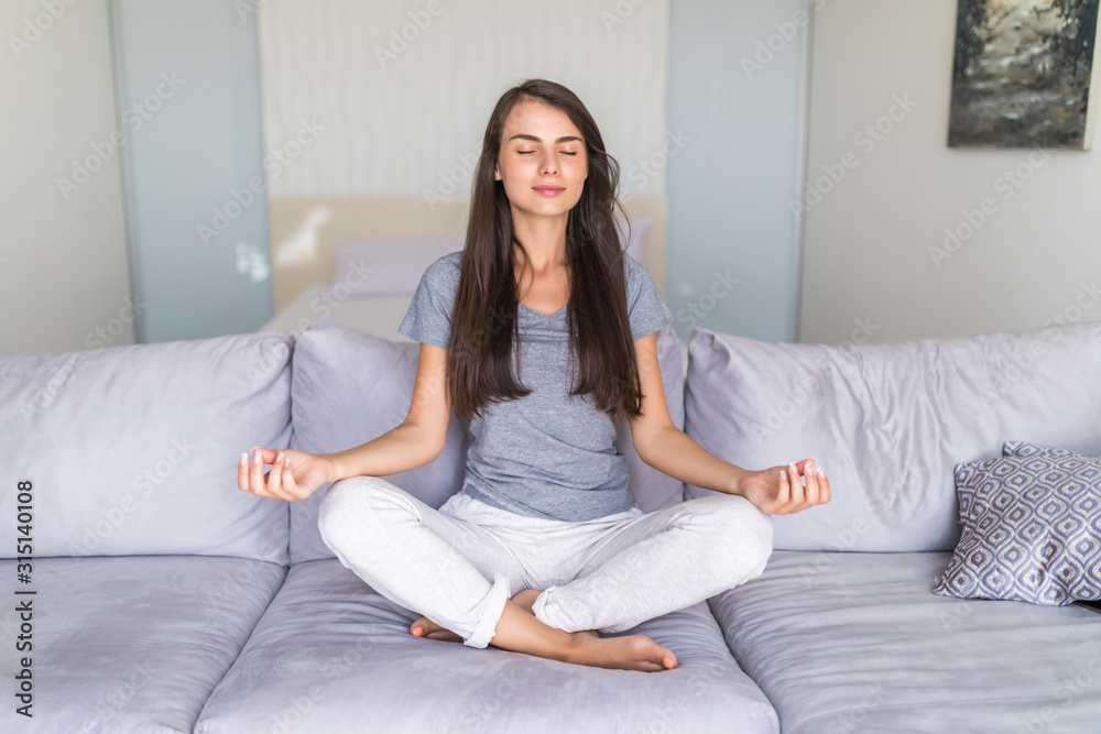 Charming young lady in casual clothes meditating on couch in lotus position, doing yoga exercises in living room in morning, relaxing and making autosuggestion practices to get positive thinking