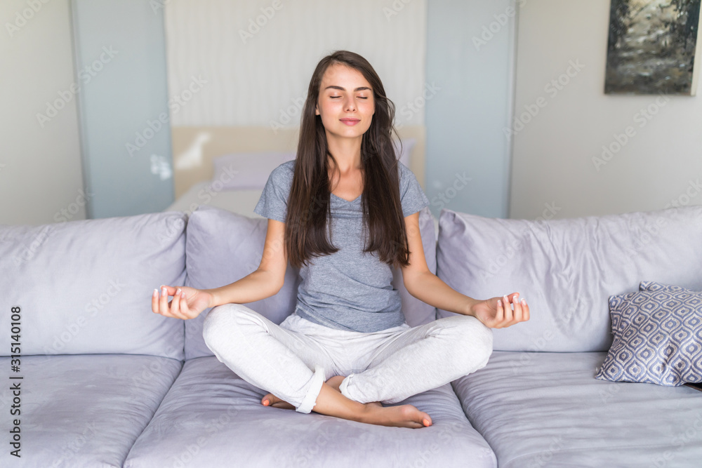 Charming young lady in casual clothes meditating on couch in lotus position, doing yoga exercises in living room in morning, relaxing and making autosuggestion practices to get positive thinking Charming young lady in casual clothes meditating on couch in lotus position, doing yoga exercises in living room in morning, relaxing and making autosuggestion practices to get positive thinking