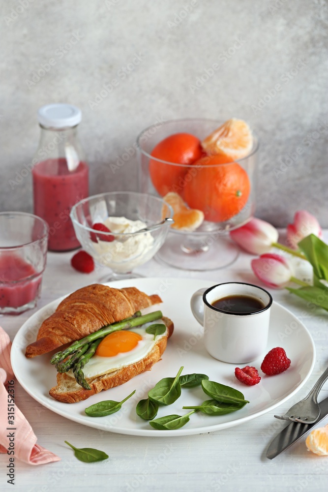  Festive breakfast table with croissant sandwich, fried egg, asparagus,  smoothie and fresh raspberry.