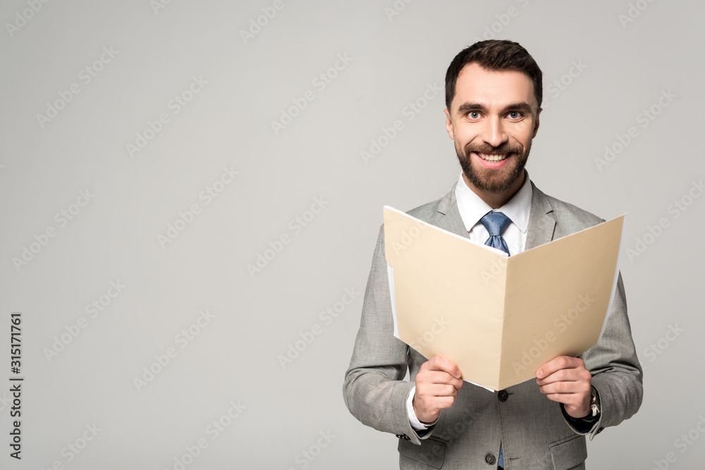 positive businessman smiling at camera while holding paper folder isolated on grey positive businessman smiling at camera while holding paper folder isolated on grey